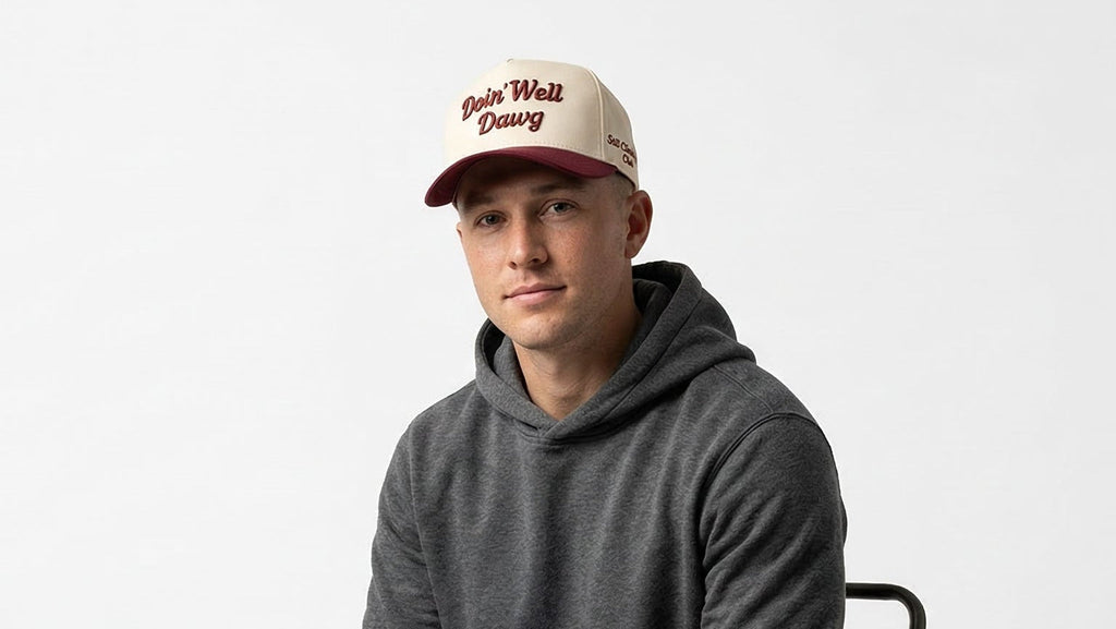 Man sitting on a chair wearing a gray hoodie and a cap with text on it against a white background