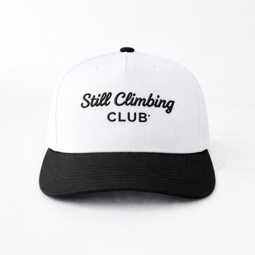 White and black cap with 'Still Climbing Club' text on a white background