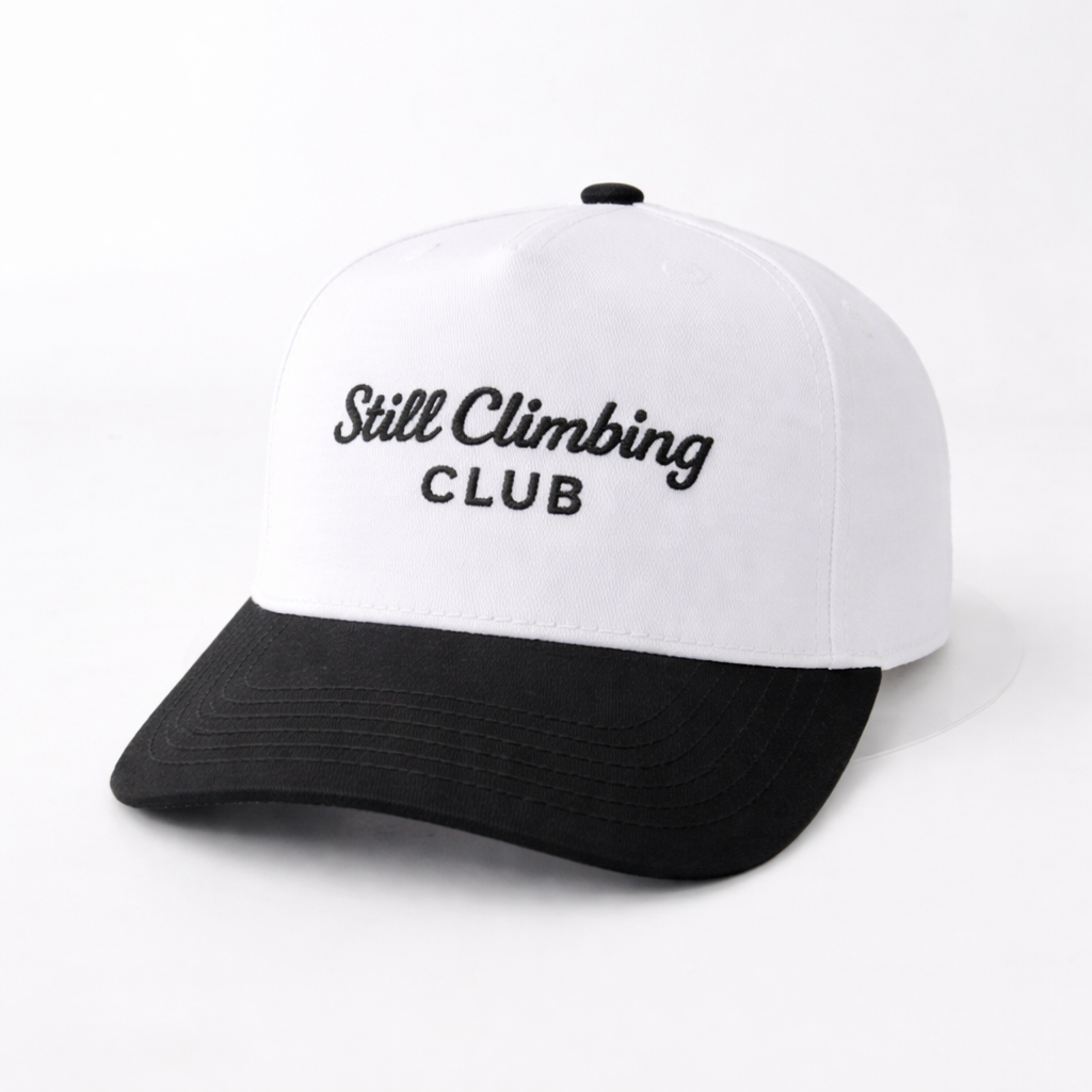 White and black cap with 'Still Climbing Club' text on a white background
